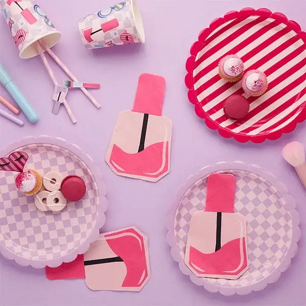 Pamper party tableware with pink nail polish shaped decorations, striped plates, cups, sweets, and pastel accessories arranged on a soft background